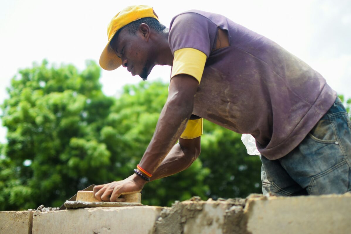 A man in a purple T-shirt and blue denim jeans utilising a brick trowel to spread mortar