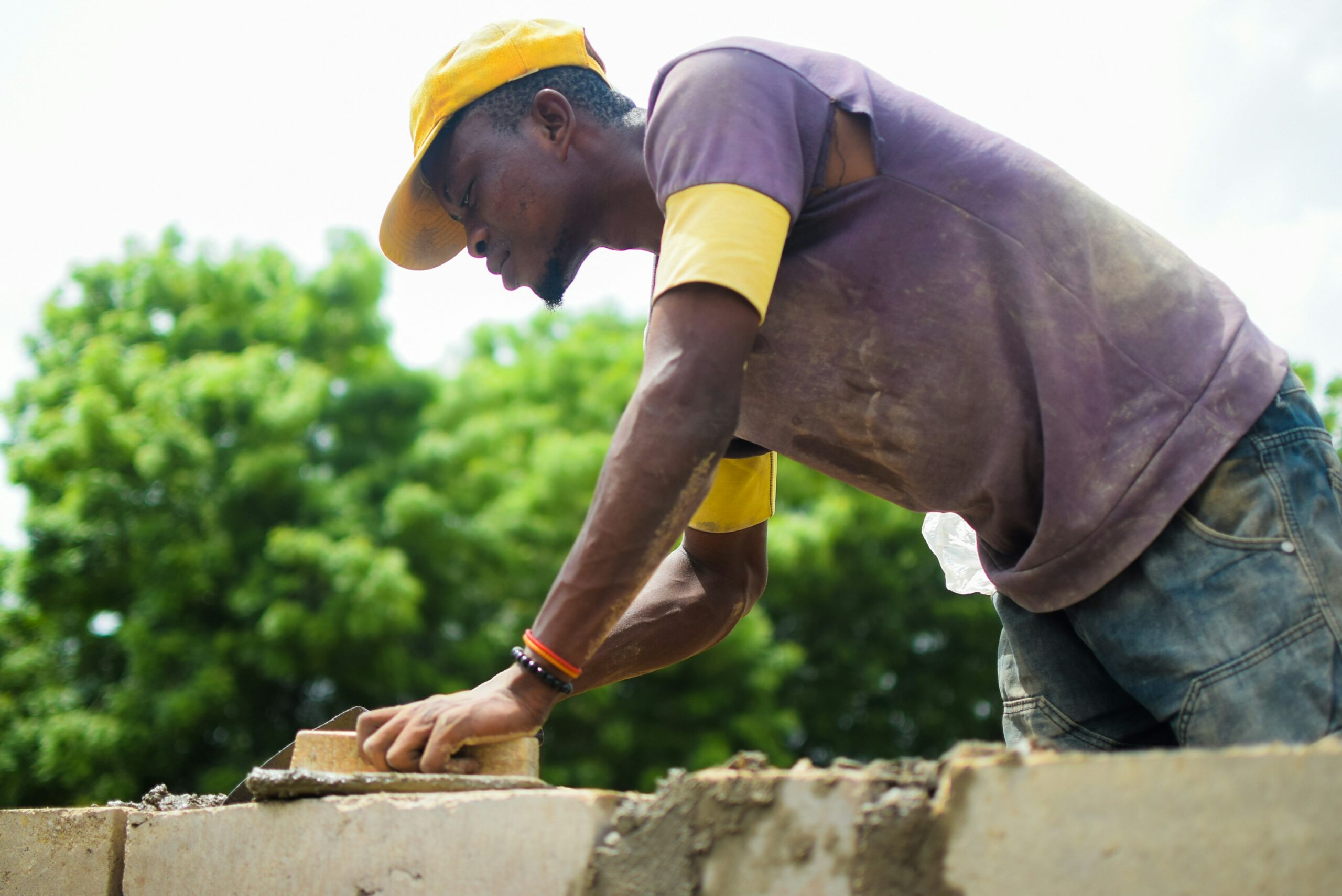 A man in a purple T-shirt and blue denim jeans utilising a brick trowel to spread mortar