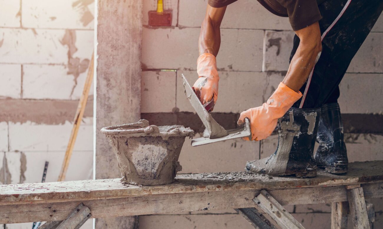 Close up of a worker wearing orange gloves standing on a wooden bench and using a brick trowel for plastering a house