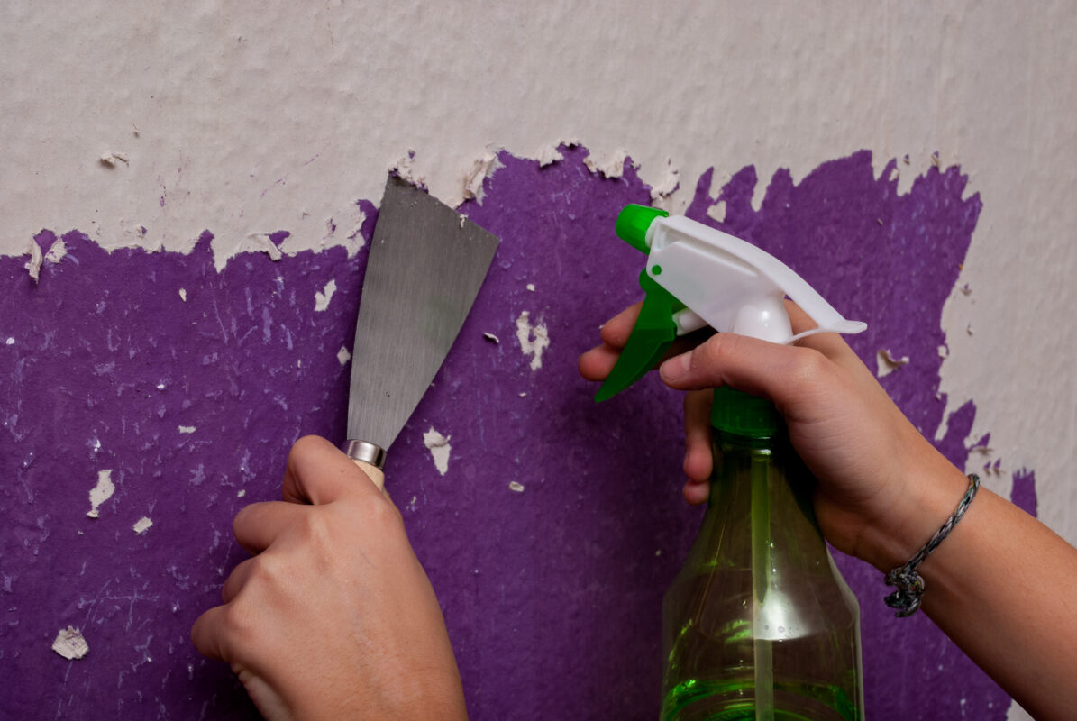 A person using a paint stripper and scraper to remove paint from a purple wall